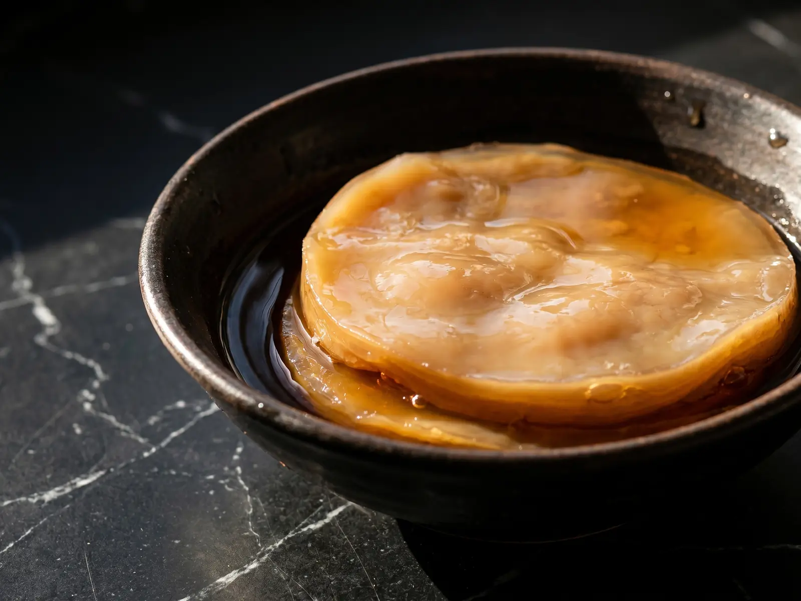 kombucha scoby Cuenco met donkere thee en een beige SCOBY op mat zwart marmer in natuurlijk licht.