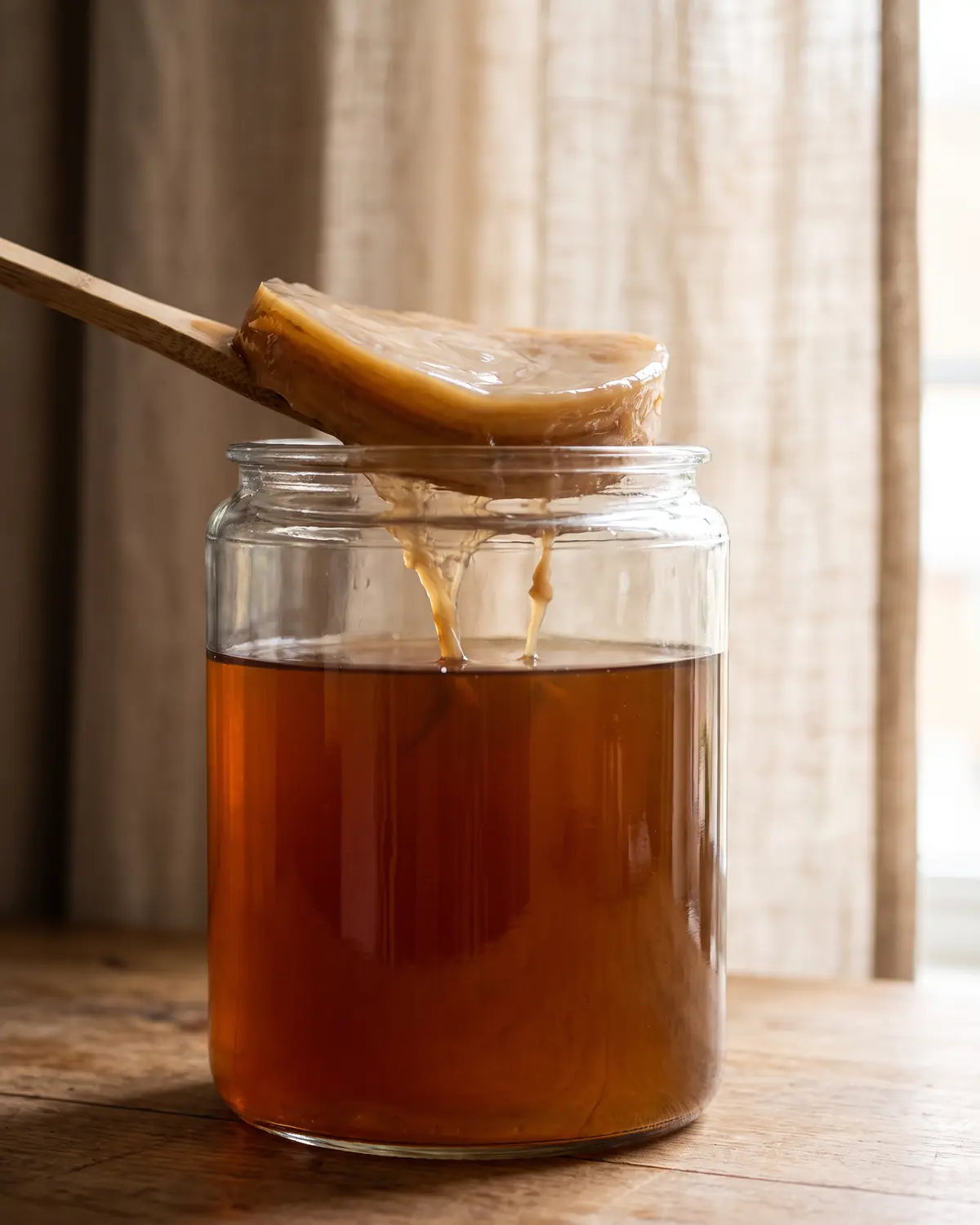 scoby hotel Glazen pot met SCOBY en zachte draden naast een onscherpe rand van een linnen gordijn.