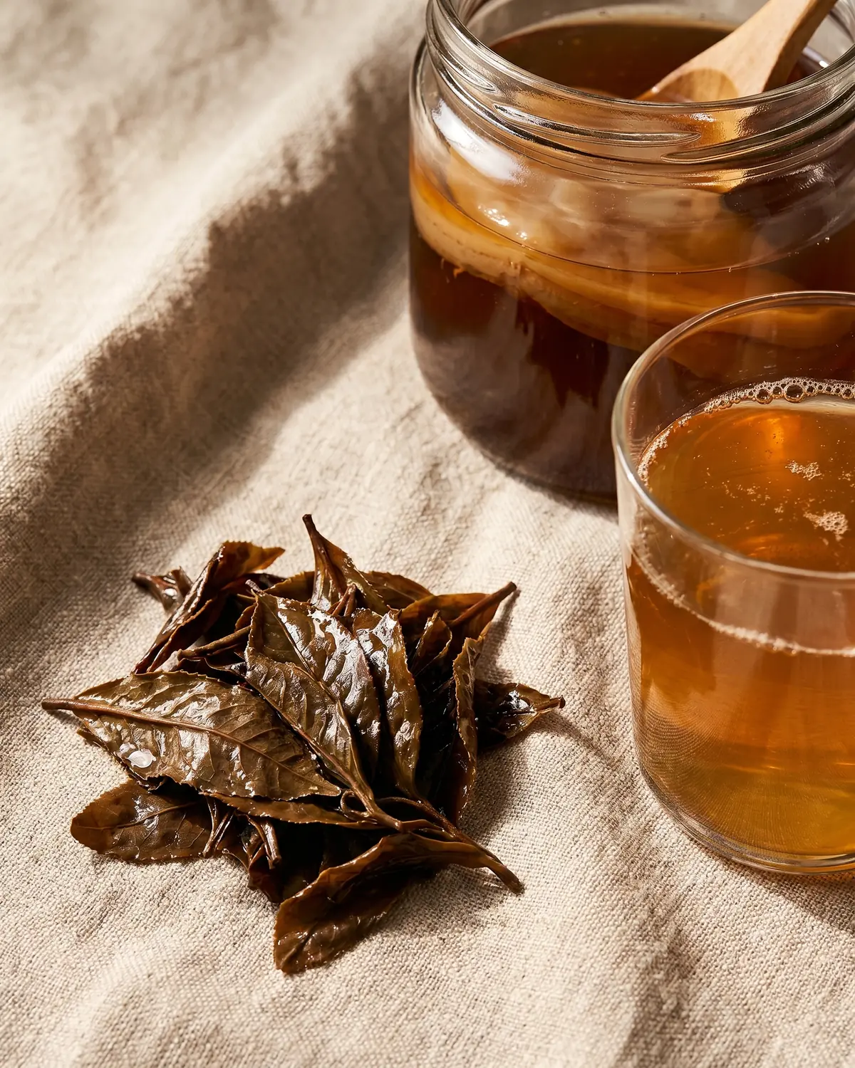 Vochtige theebladeren naast een glas kombucha op ruw linnen, als detail van het fermentatieproces.