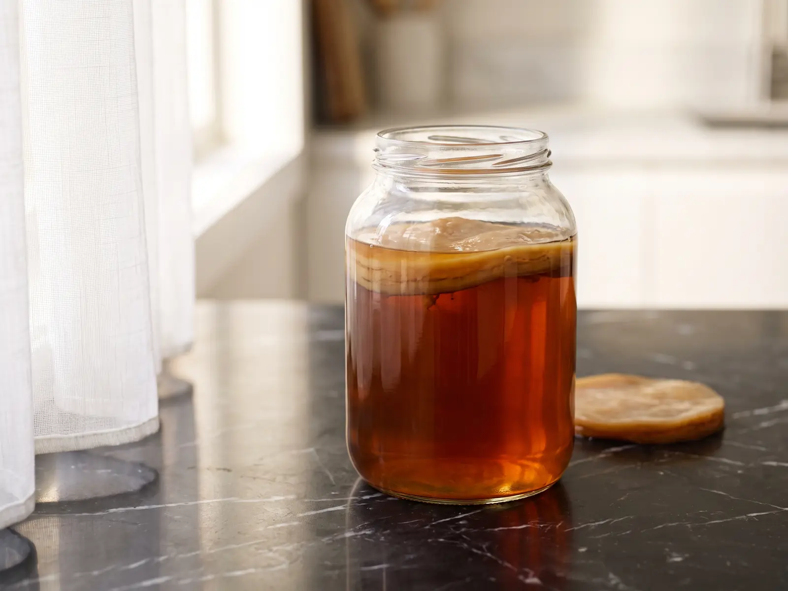 kombucha benefits Pot met SCOBY en amberkleurige kombucha op zwart marmer met zachte spiegeling van een wit gordijn
