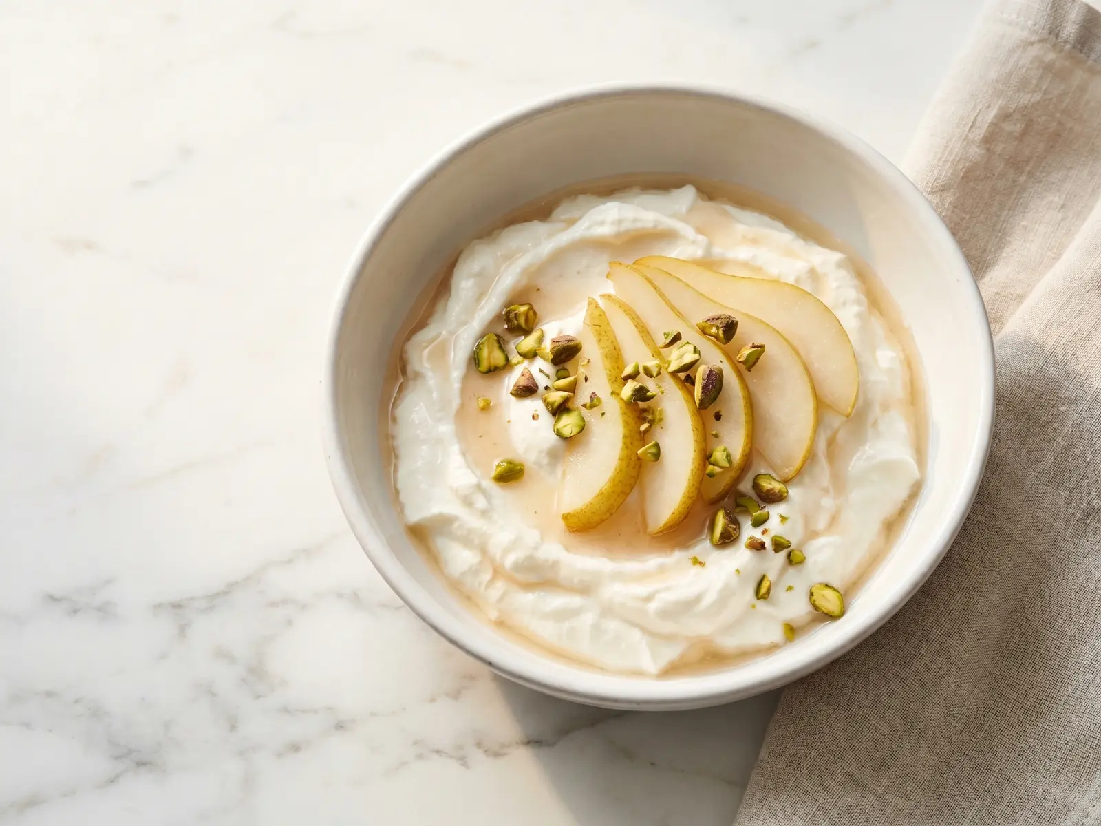 Romige yoghurt in een witte keramieken kom met plakjes peer en pistachenoten op geaderd marmer.